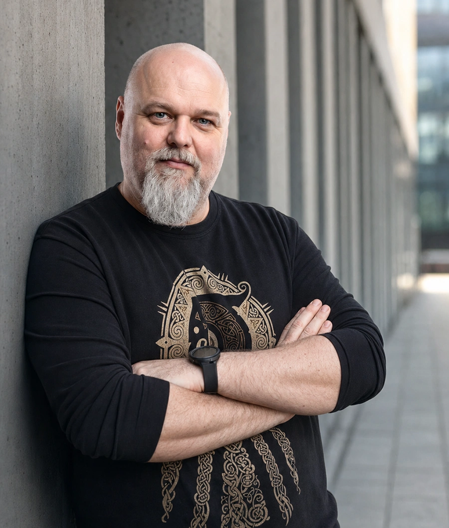 Pewny siebie mężczyzna w czarnej koszulce z zegarkiem na tle nowoczesnego budynku Uśmiechnięty mężczyzna z brodą oparty o betonową ścianę, ubrany w czarną koszulkę z wzorem