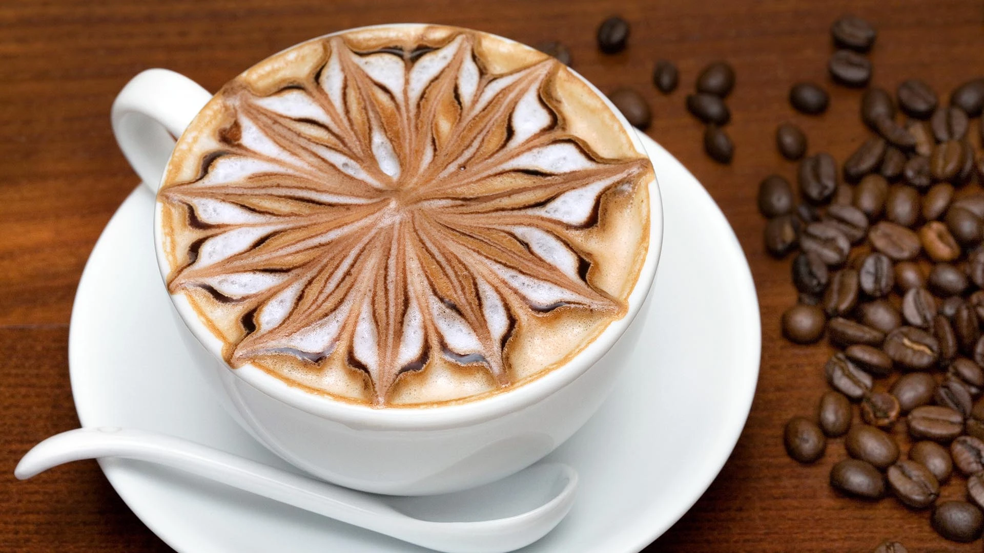 Filiżanka cappuccino z artystycznym wzorem na mlecznej piance, obok rozsypane ziarna kawy.