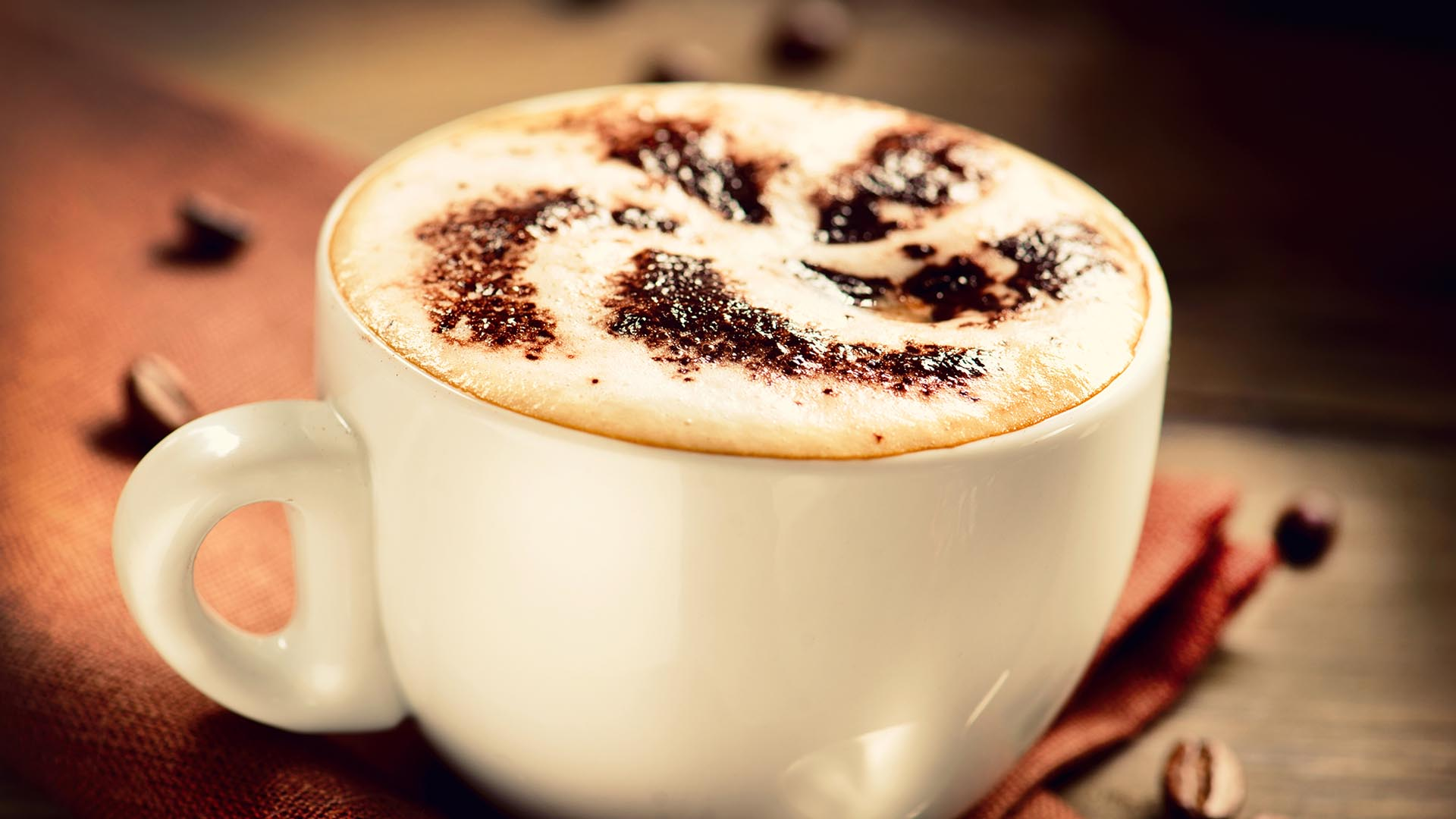 Kawa cappuccino z pianką i posypką w białej filiżance na tle brązowej serwetki.