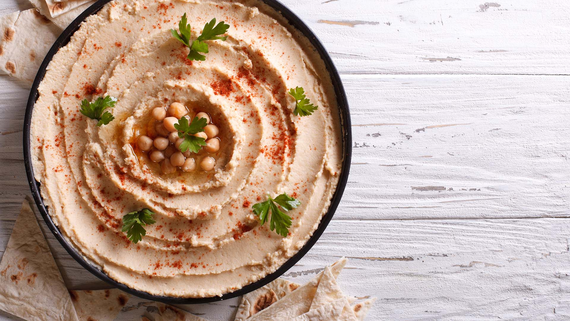 Kremowy hummus z ciecierzycą i papryką w czarnej misce, udekorowany natką pietruszki na białym stole.