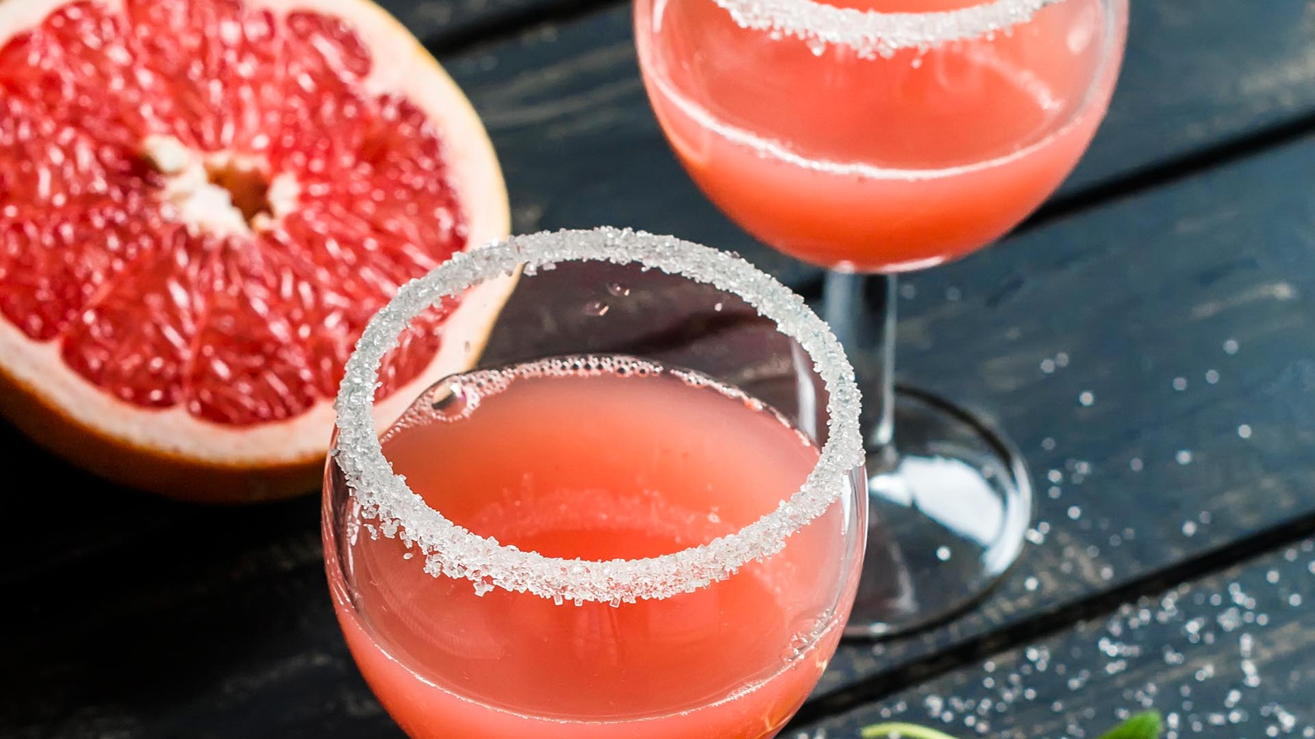 Koktajl grapefruitowy w kieliszku z cukrową obwódką na tle połówki owocu.