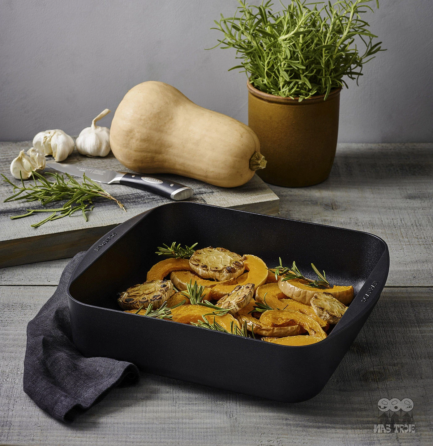 Kwadratowa patelnia nieprzywierająca Scanpan z warzywami i ziołami, idealna do serwowania. Tytanowo-ceramiczna patelnia Scanpan z pieczonymi warzywami i ziołami na tle kuchennego blatu.