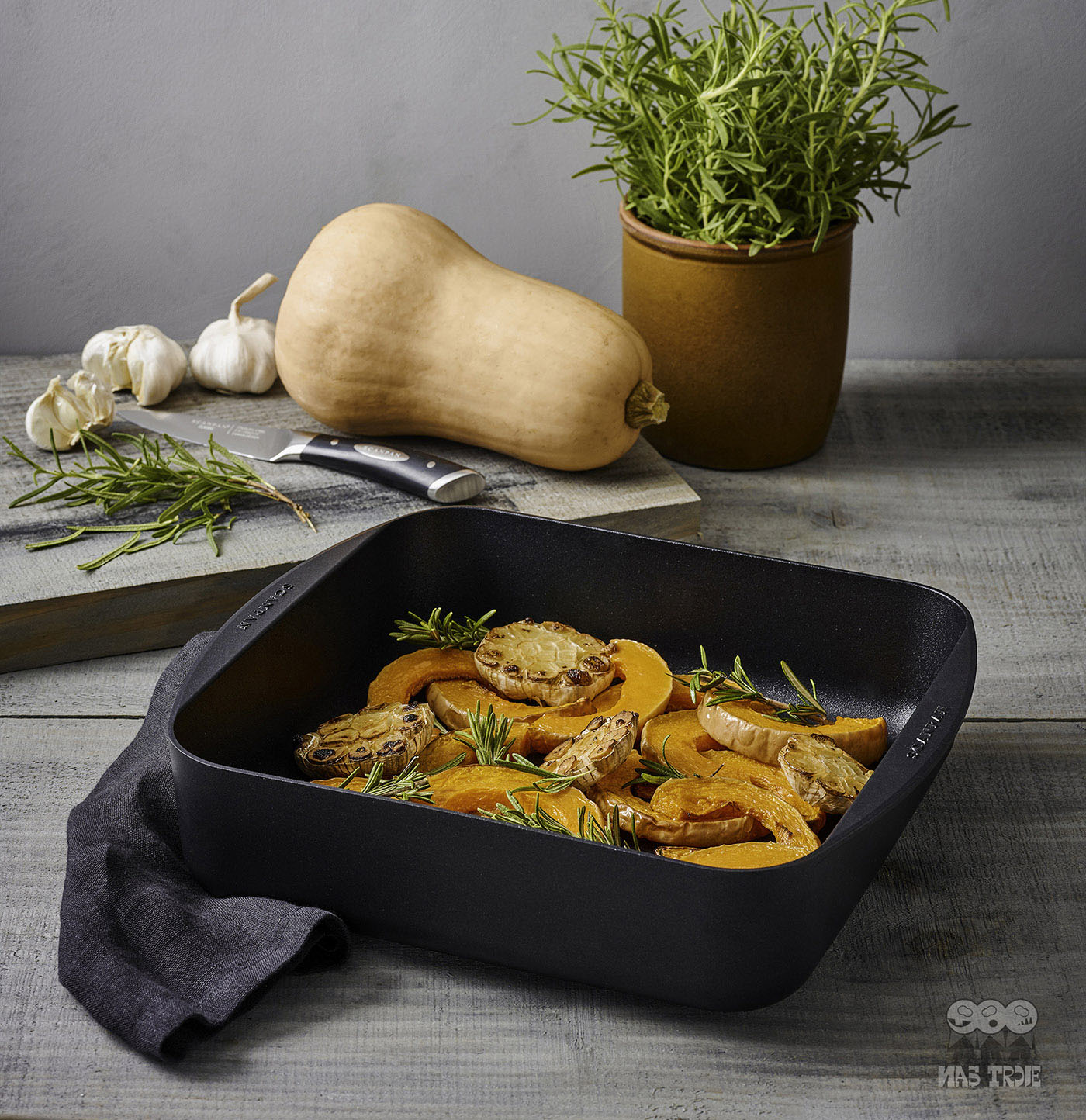 Kwadratowa patelnia nieprzywierająca Scanpan z warzywami i ziołami, idealna do serwowania. Tytanowo-ceramiczna patelnia Scanpan z pieczonymi warzywami i ziołami na tle kuchennego blatu.