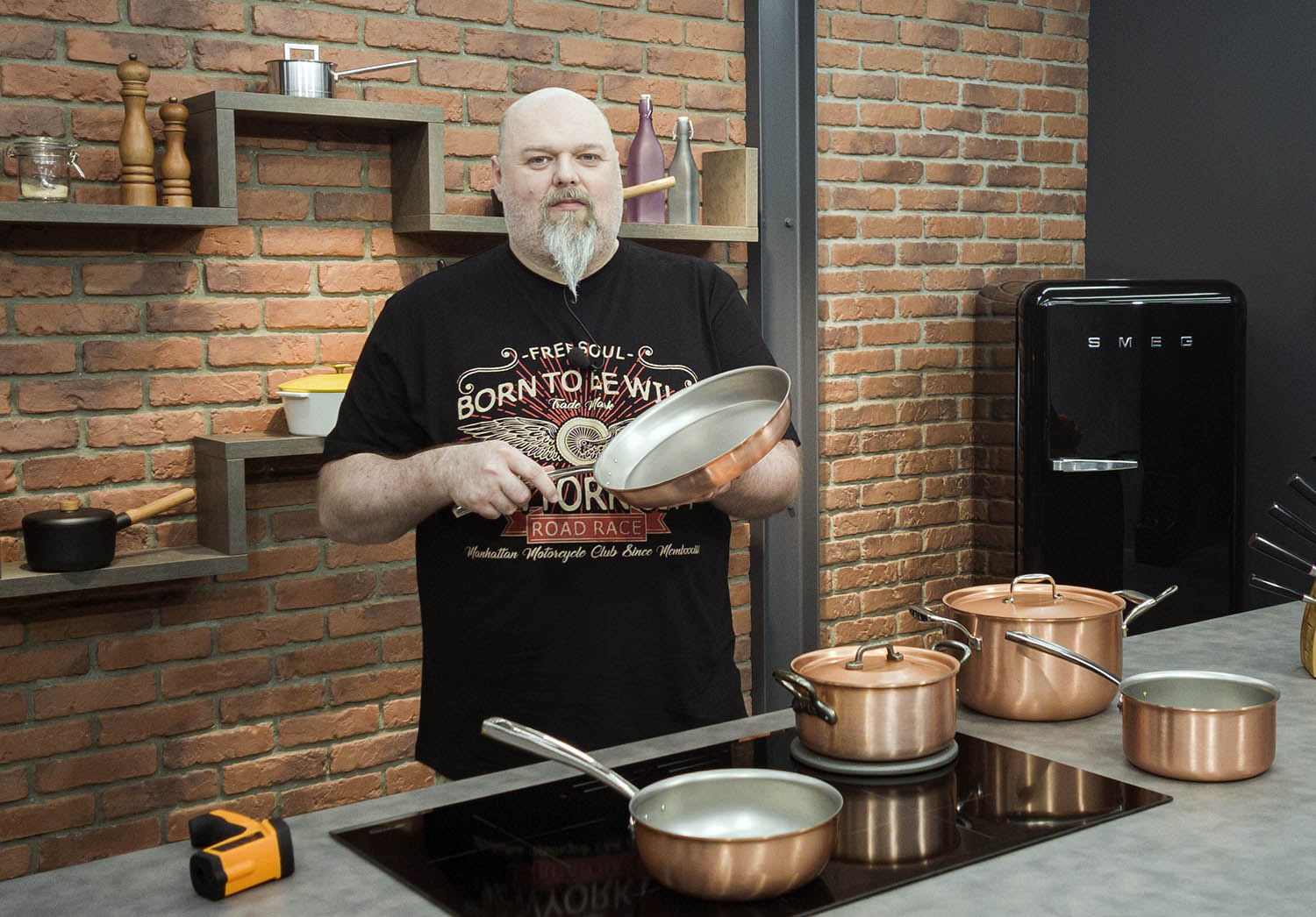 Miedziana patelnia Falk w kuchni z ceglaną ścianą, trzymana przez osobę w czarnej koszulce.