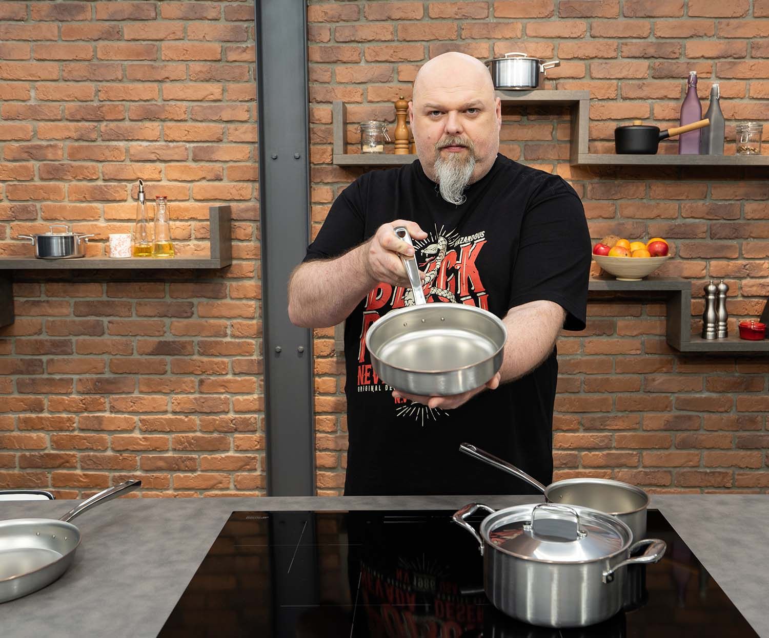 Mężczyzna prezentuje stalowo-miedzianą patelnię sauté w kuchni z ceglaną ścianą.