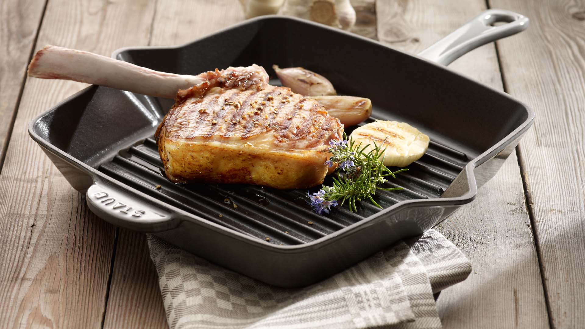 Żeliwna patelnia grillowa Staub z mięsem i ziołami na drewnianym stole.