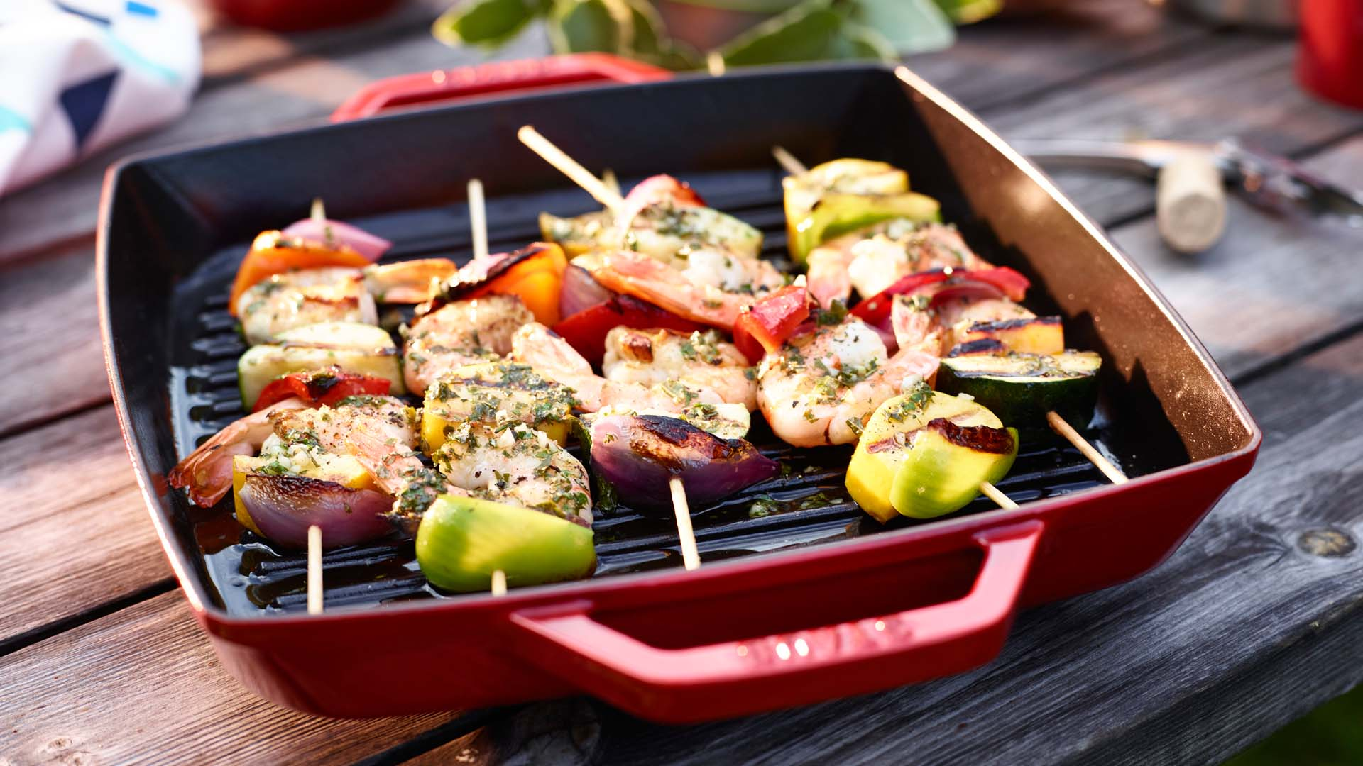 Szaszłyki na żeliwnej patelni Staub, kwadratowy model do grillowania, na tle drewnianego stołu. Kwadratowa żeliwna patelnia grillowa Staub z szaszłykami warzywno-mięsnymi na drewnianym stole.