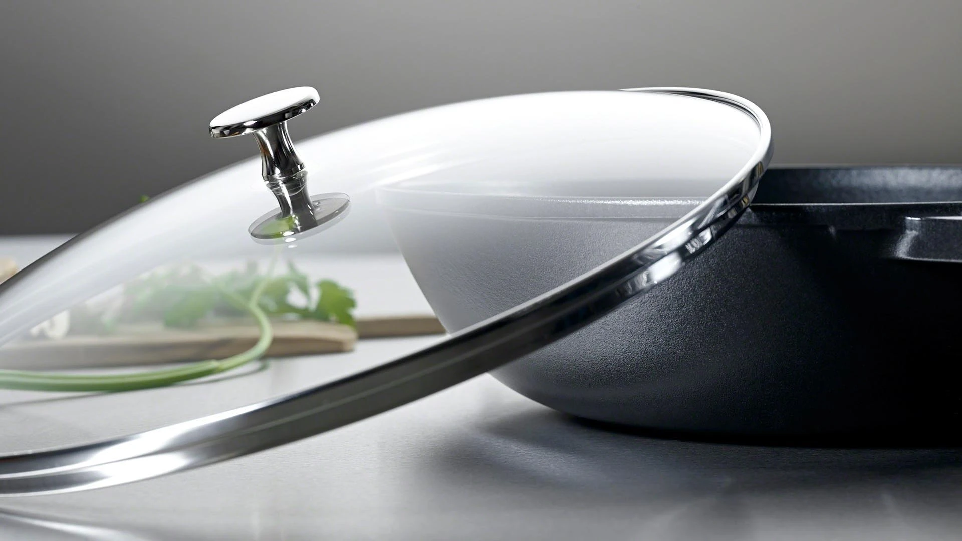 Wok Staub z nieprzywierającą powłoką i szklaną pokrywą. Żeliwny wok z przezroczystą pokrywą Staub na tle kuchennego blatu.