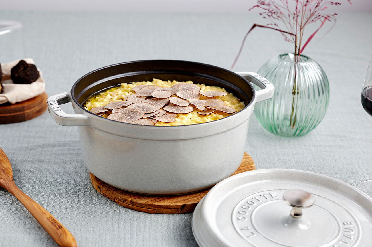 Owalny żeliwny garnek Staub z białymi truflami na risotto, stojący na drewnianej podstawce.