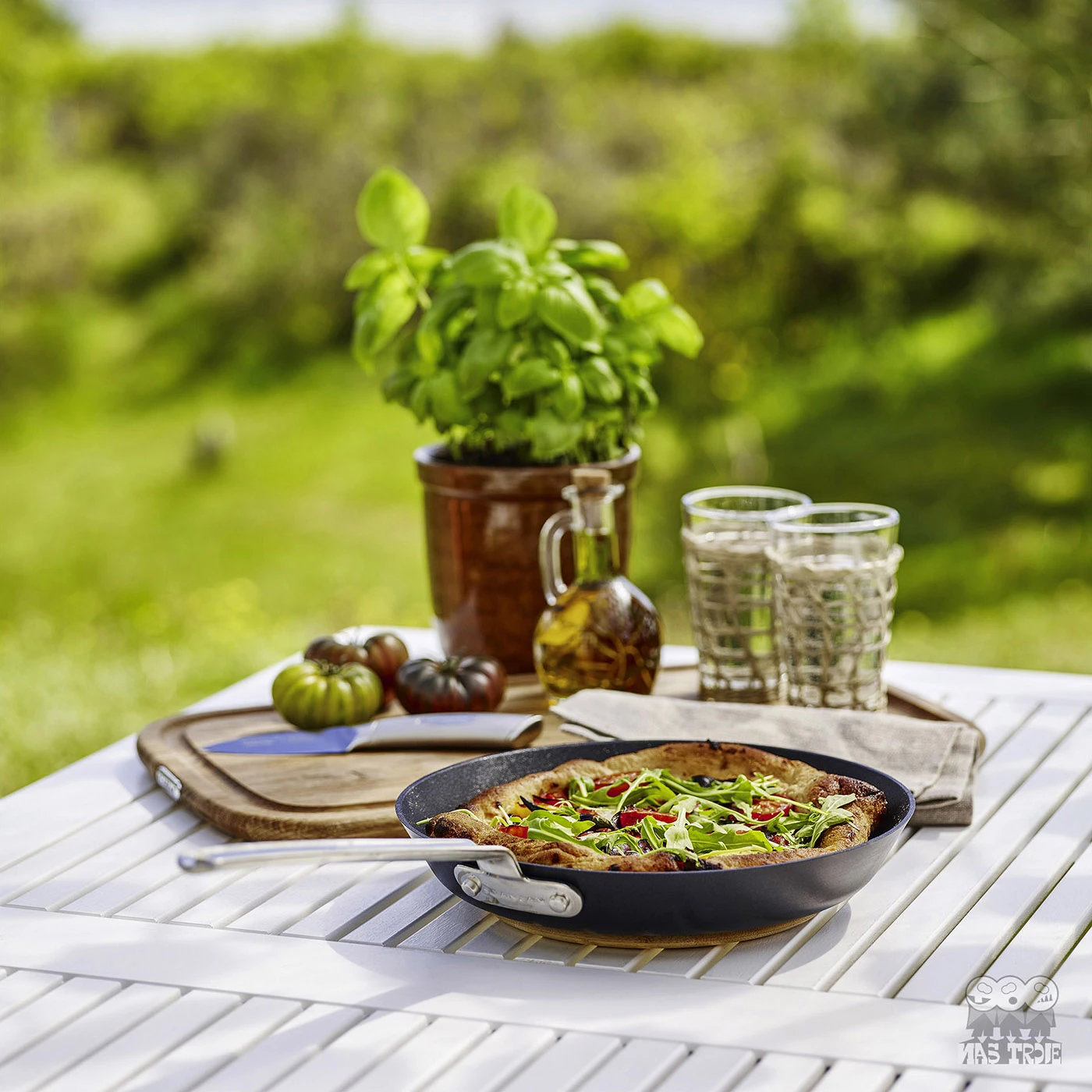 Scanpan TechnIQ z pizzą na tarasie, obok świeżych ziół i napojów. Patelnia tytanowo-ceramiczna z pizzą na białym stole w ogrodzie, obok ziół i szklanek.
