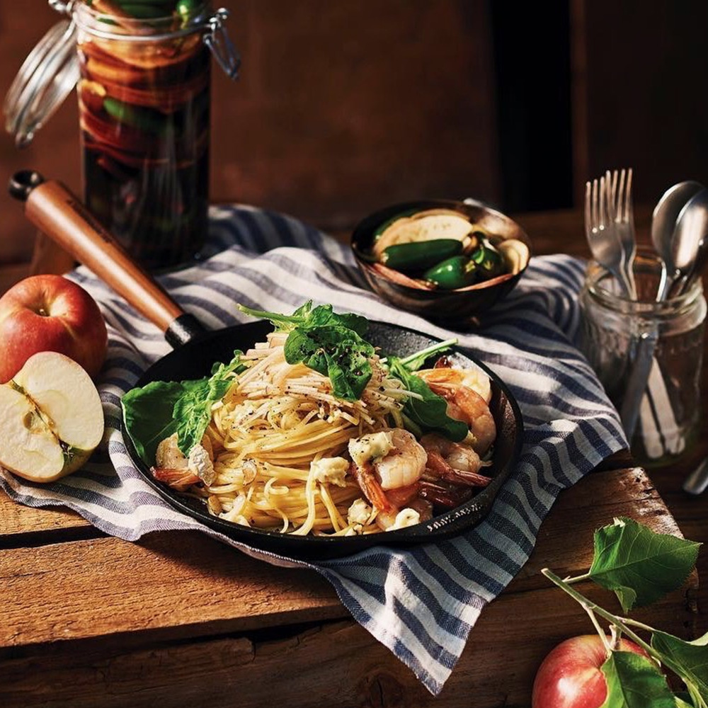 Płytka patelnia z żeliwa Skeppshult, serwująca spaghetti z krewetkami i świeżymi dodatkami. Żeliwna patelnia Skeppshult z orzechowym uchwytem, pełna spaghetti z krewetkami i ziołami.