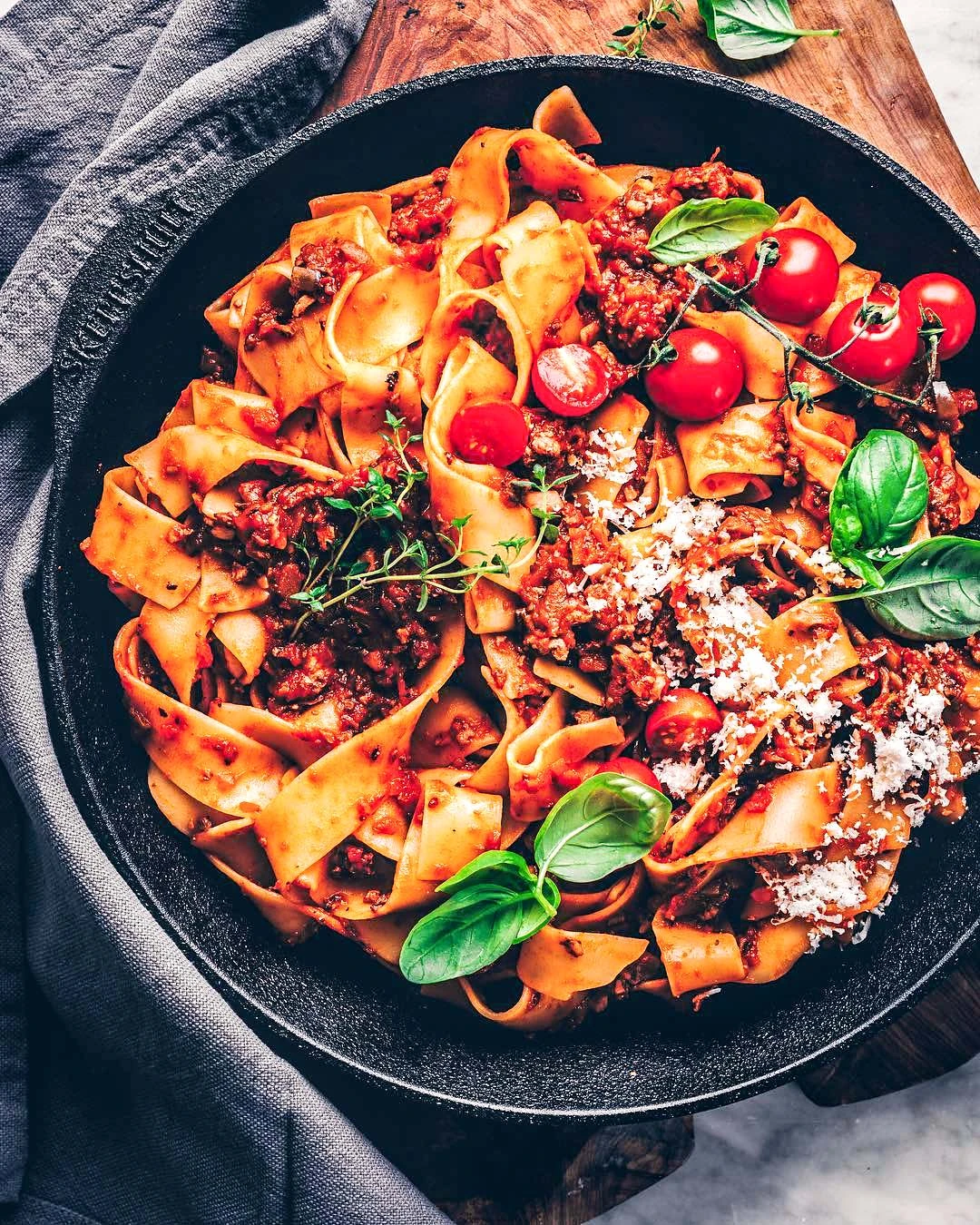 Żeliwny wok z makaronem pappardelle, pomidorkami i bazylią na drewnianej desce.