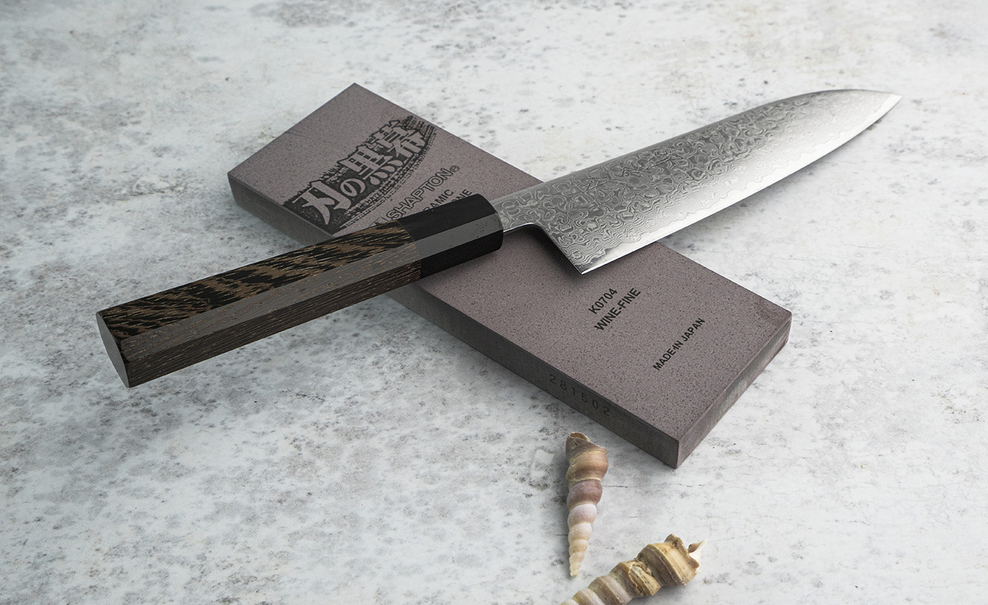 Japoński nóż Santoku 18 cm z 69-warstwowej stali, rękojeść z drewna, na tle kamienia ostrzącego.