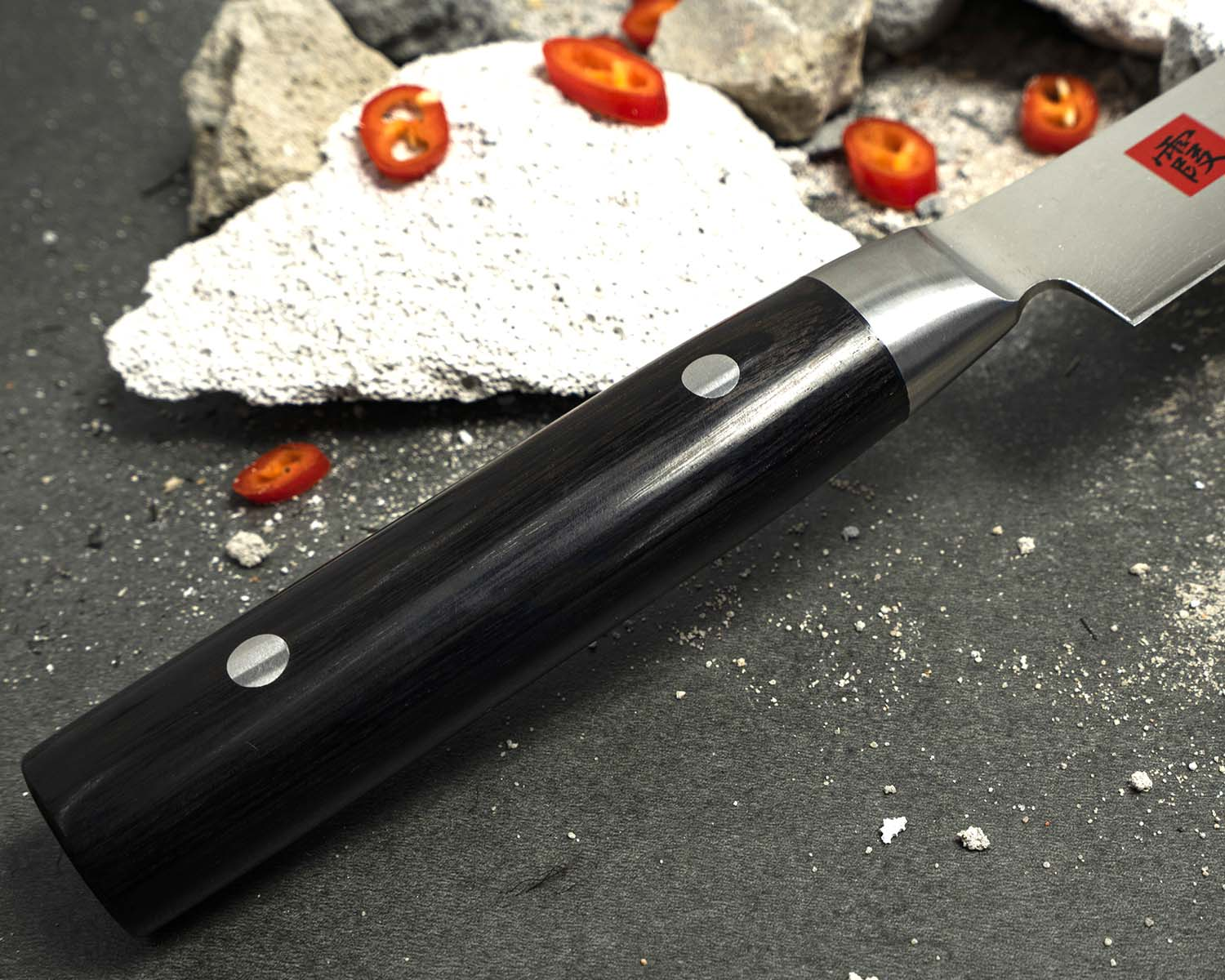 Elegancki nóż Santoku z ostrzem z damasceńskiej stali i drewnianą rękojeścią. Japoński nóż Santoku z rękojeścią z ciemnego drewna na tle pokruszonego kamienia i plasterków papryki.