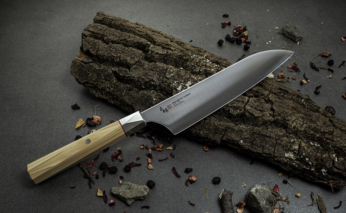 Japoński nóż Santoku Mcusta Zanmai na tle kory drzewa i suszonych ziół.