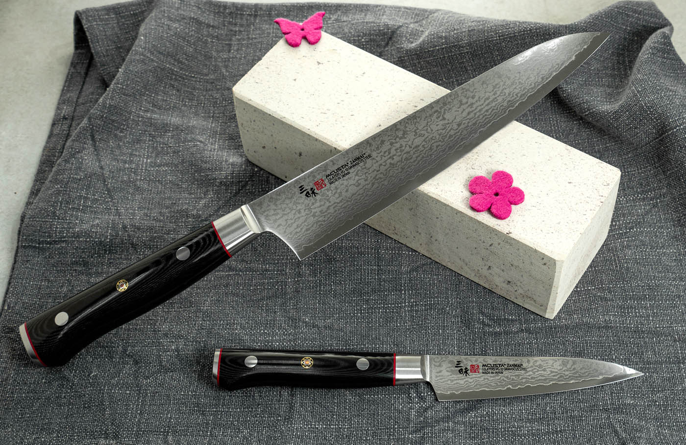 Profesjonalny nóż sushi Mcusta Zanmai z 33-warstwową klingą na ciemnym tle. Japoński nóż Sujihiki 27 cm Mcusta Zanmai na tle kamienia i szarej tkaniny.