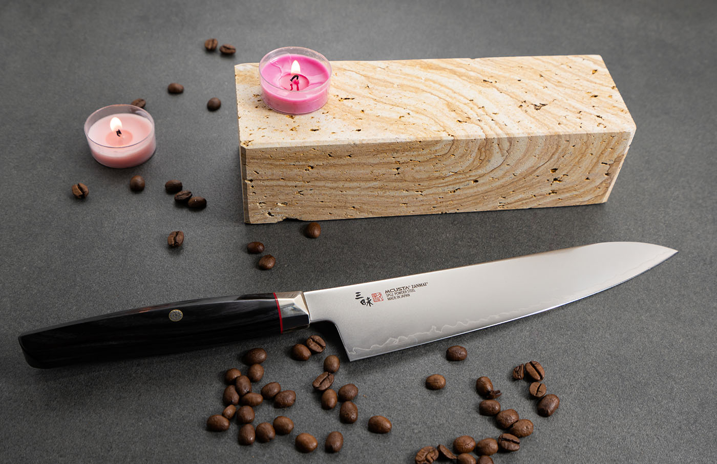 Japoński nóż Mcusta Zanmai Ko-Santoku z ozdobnym tłem. Nóż Ko-Santoku Mcusta Zanmai z różowymi świecami na tle kamiennego bloku.