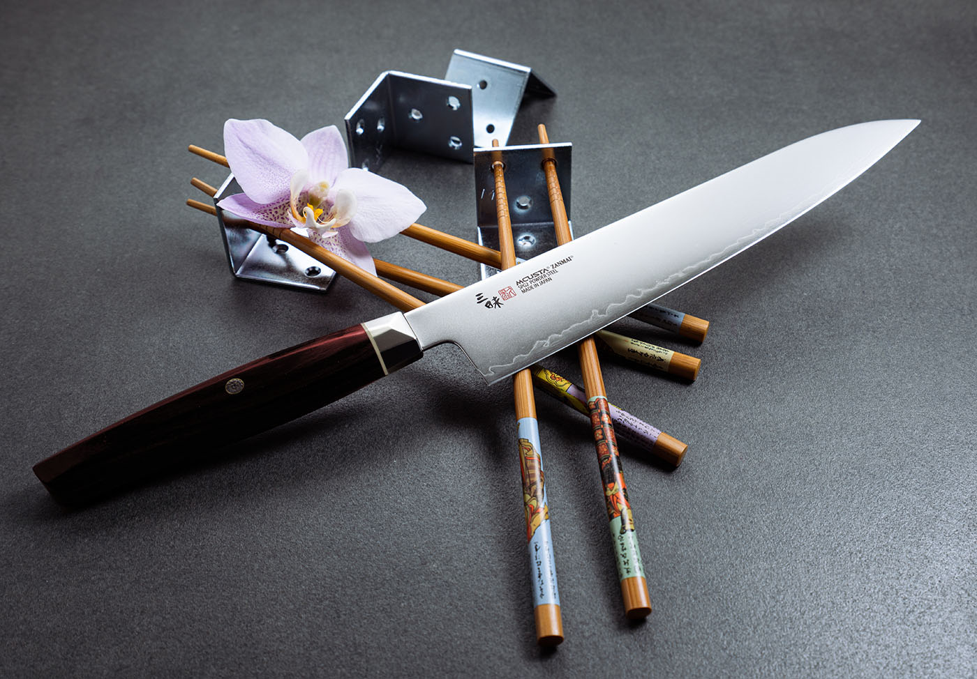 Elegancki nóż Ko-Santoku Mcusta Zanmai, stal SPG2, w otoczeniu pałeczek i kwiatów. Japoński nóż Ko-Santoku z rękojeścią z ciemnego drewna na tle pałeczek i orchidei.