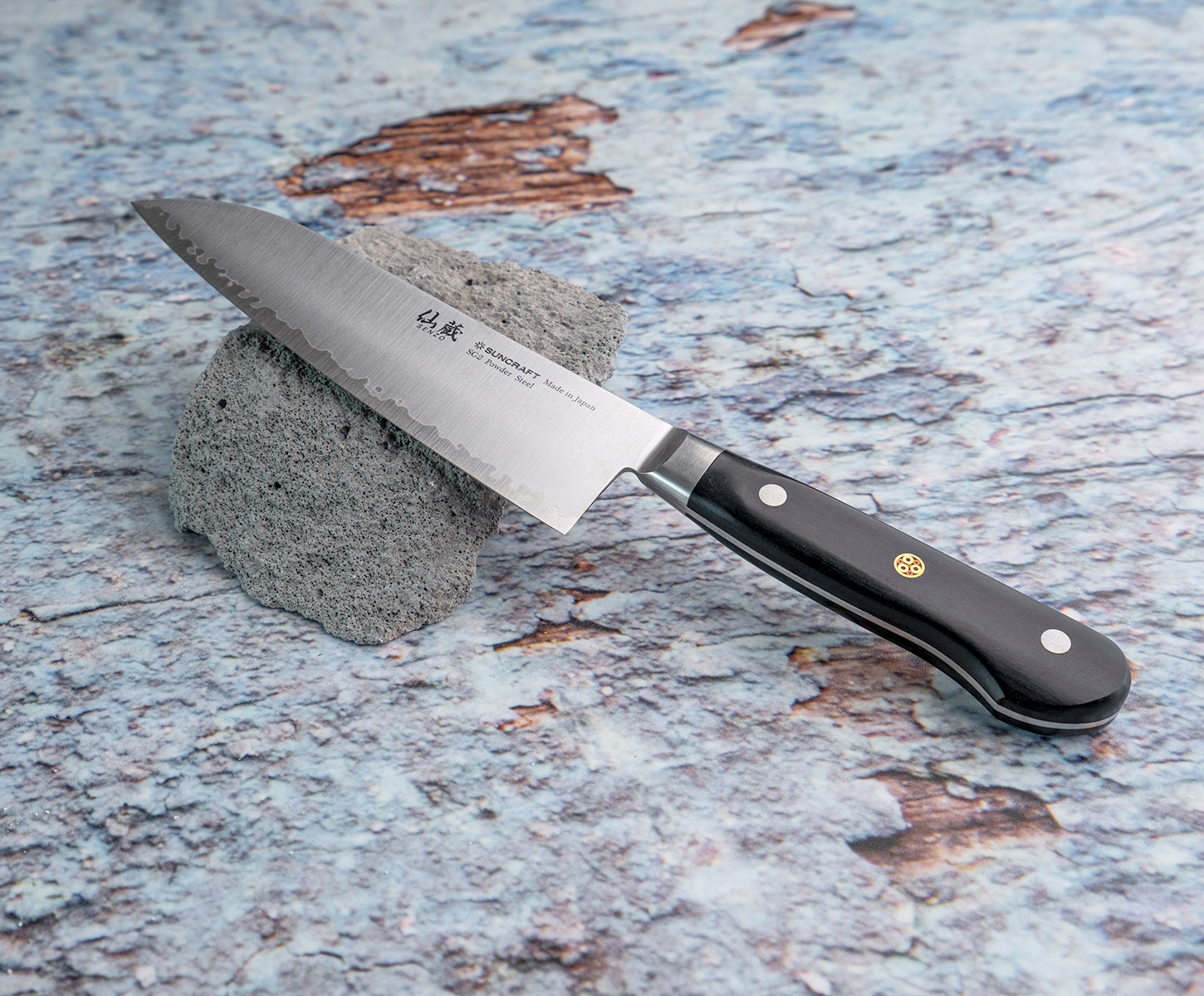 Japoński nóż Santoku Suncraft z czarną rękojeścią, oparty na kamieniu.