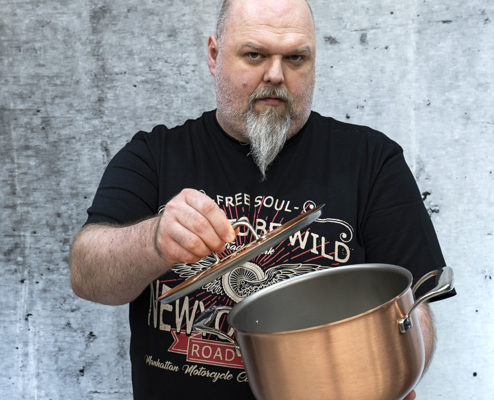 Mężczyzna prezentuje niski garnek miedziany AvantGarde z pokrywką. Miedziany garnek AvantGarde z pokrywką i stalowymi uchwytami, trzymany przez mężczyznę.