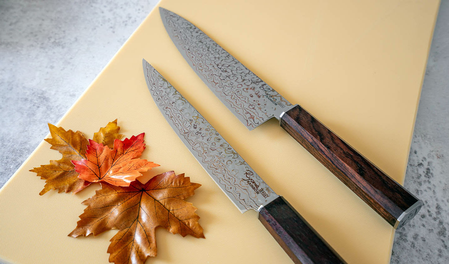 Nóż Santoku Ryoma Rainbow z damasceńskiej stali, ozdobiony liśćmi na stole. Japoński nóż Santoku Ryoma Rainbow na tle deski do krojenia i jesiennych liści.