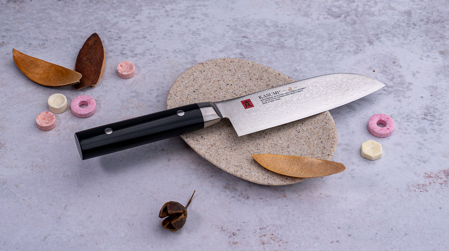Japoński nóż Santoku z damasceńskiej stali VG-10 na kamiennej powierzchni, ozdobiony liśćmi i kolorowymi pastylkami.