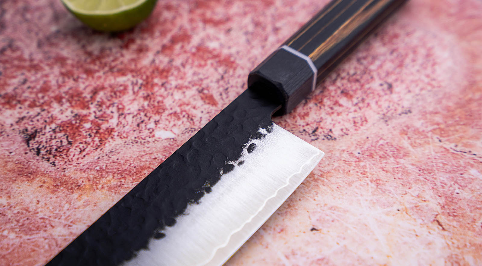 Japoński nóż Santoku 17 cm z czarną rękojeścią i ostrzem na tle różowego blatu.
