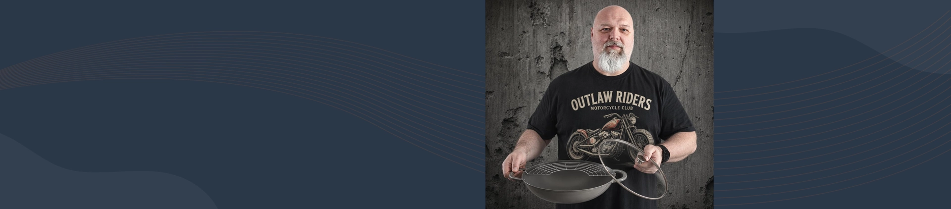 Mężczyzna trzyma żeliwny wok Staub Graphite Grey z rusztem, na tle betonowej ściany.