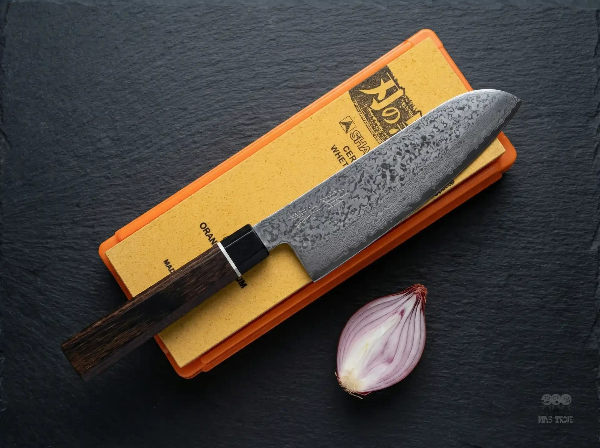 Japoński nóż Santoku Senzo Black na osełce ceramicznej obok przekrojonej czerwonej szalotki.