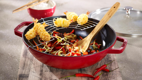 Wok żeliwny Staub Graphite Grey z warzywami i krewetkami na drewnianej desce.
