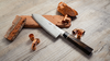 Japoński nóż Santoku Suncraft Senzo Black na tle cegieł i miedzianych elementów.