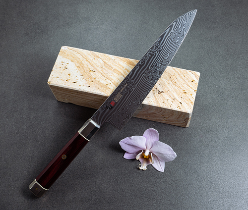 Nóż Santoku Mcusta Zanmai Ultimate Aranami na tle kamienia i kwiatu.