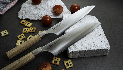 Japoński nóż Santoku Mcusta Zanmai Beyond, stal Aogami Super, 18 cm, w otoczeniu przekąsek i pomidorów.