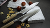 Japoński nóż Santoku Mcusta Zanmai Beyond, stal Aogami Super, 18 cm, w otoczeniu przekąsek i pomidorów.