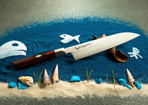 Młotkowany nóż Gyuto 24 cm Mcusta Zanmai Supreme Ripple na tle dekoracji morskiej.