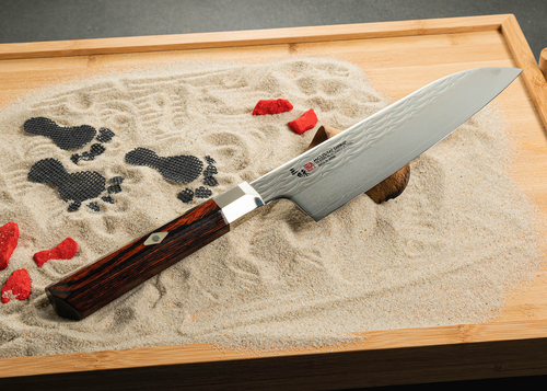 Nóż Santoku Mcusta Zanmai Supreme Ripple na piaskowym tle.