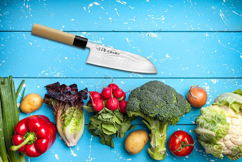 Japoński nóż Santoku Tojiro Shippu na tle świeżych warzyw na niebieskim stole.