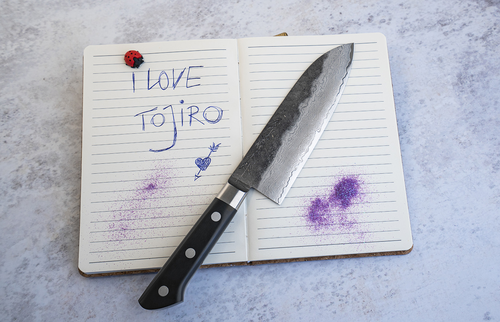 Japoński nóż Santoku Hatsukokoro Atelier, mistrz Tomoo Matsumura, na tle zeszytu.