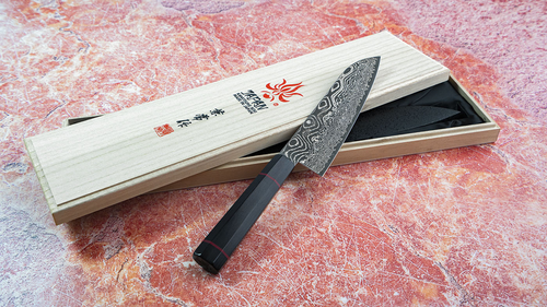 Japoński nóż Santoku Kanetsune Namishibuki w drewnianym pudełku na tle czerwonego kamienia.