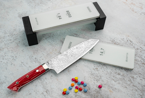 Damasceński nóż Santoku Takeshi Saji Red Turquoise 18 cm na tle ostrzałki.