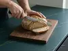 Nóż do chleba producenta produkt krojący bochenek na desce kuchennej, świeżo wypieczony chleb i dłonie.