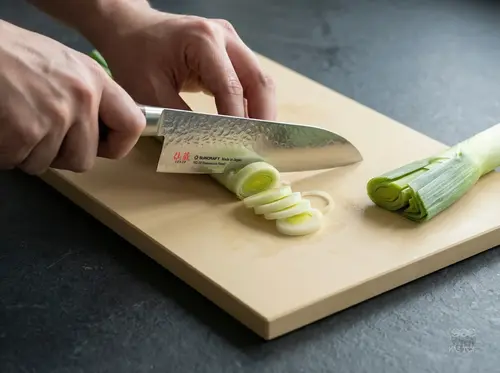 Suncraft Senzo Universal Santoku 33-warstwowy nóż japoński z VG-10 na desce podczas krojenia pora